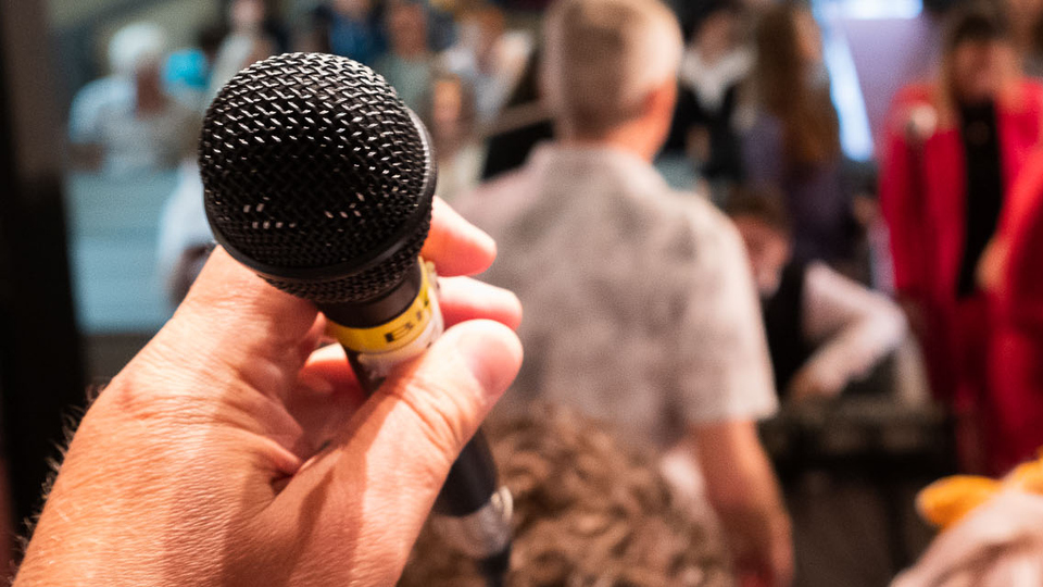 Foto från en tidigare skolavslutning som visar en hand som håller en mikrofon i bakgrunden publik och körsångare